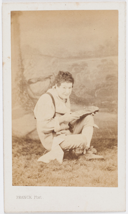Franck - Paul Legrand en habit de scène (paysan), assis les jambes croisées, ten...
