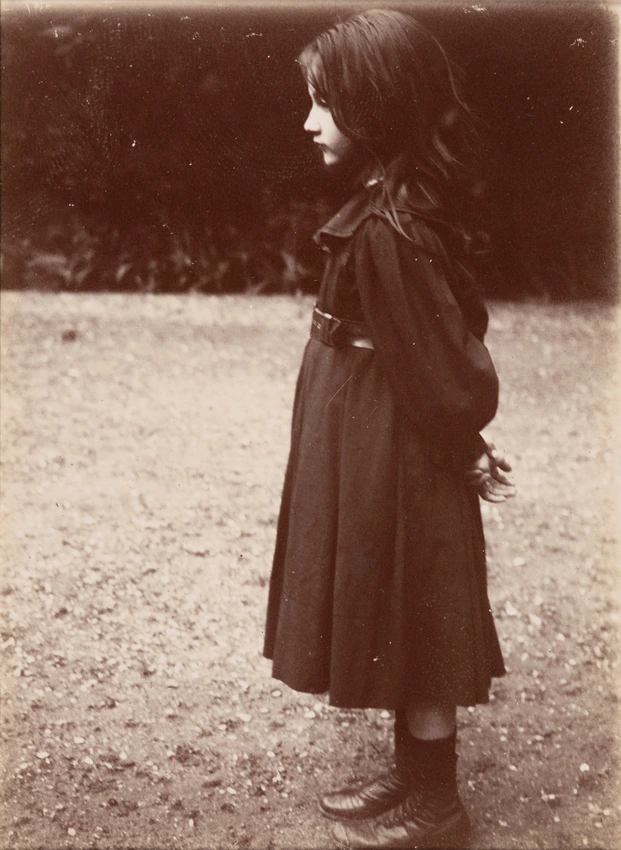 Denise debout, de profil, mains derrière elle, dans le jardin de Verneuil. Elle a les cheveux dénoués et porte une robe sombre, une ceinture et des bottines à boutons - Emile Zola