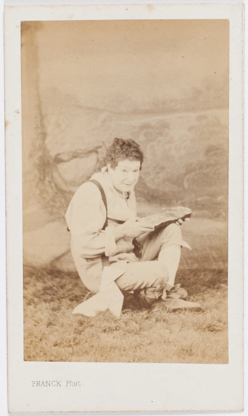 Paul Legrand en habit de scène (paysan), assis les jambes croisées, tenant une tartine de pain, une cocotte en papier contre la jambe - Franck