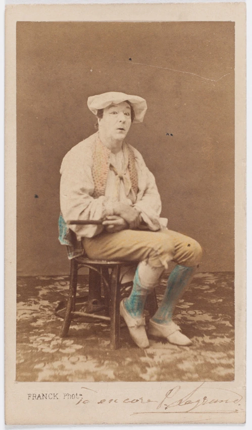 Paul Legrand en habit de scène (paysan), assis les jambes croisées, tenant une tartine de pain, une cocotte en papier contre la jambe - Franck
