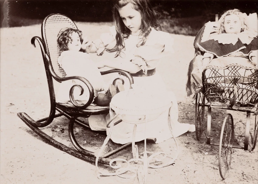 À gauche, une poupée est assise dans un fauteuil d’enfant, à bascule. Accroupie à côté du fauteuil, Denise caresse la joue de la poupée. À droite, une autre poupée est allongée dans un chariot à trois roues - Emile Zola