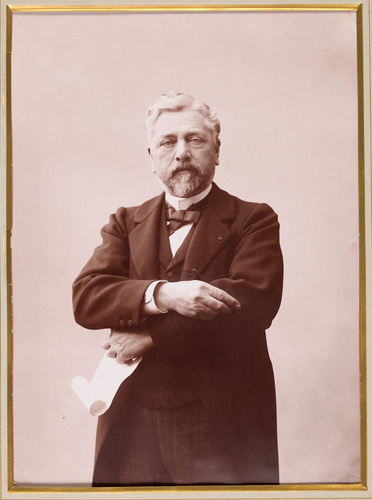 Anonyme - Gustave Eiffel debout tenant un crayon et un rouleau de papier.