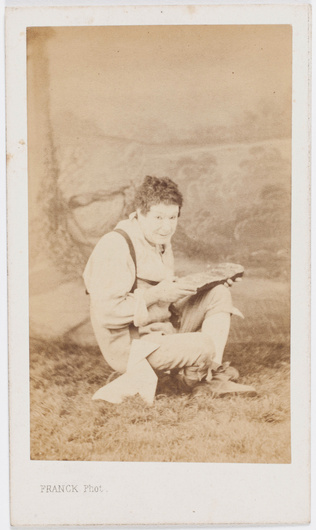 Franck - Paul Legrand en habit de scène (paysan), assis les jambes croisées, ten...