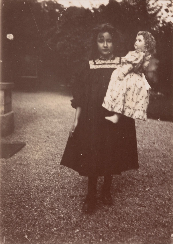 Dans une allée en graviers, Denise, au premier plan, cheveux dénoués, tient avec son bras gauche une grande poupée - Emile Zola