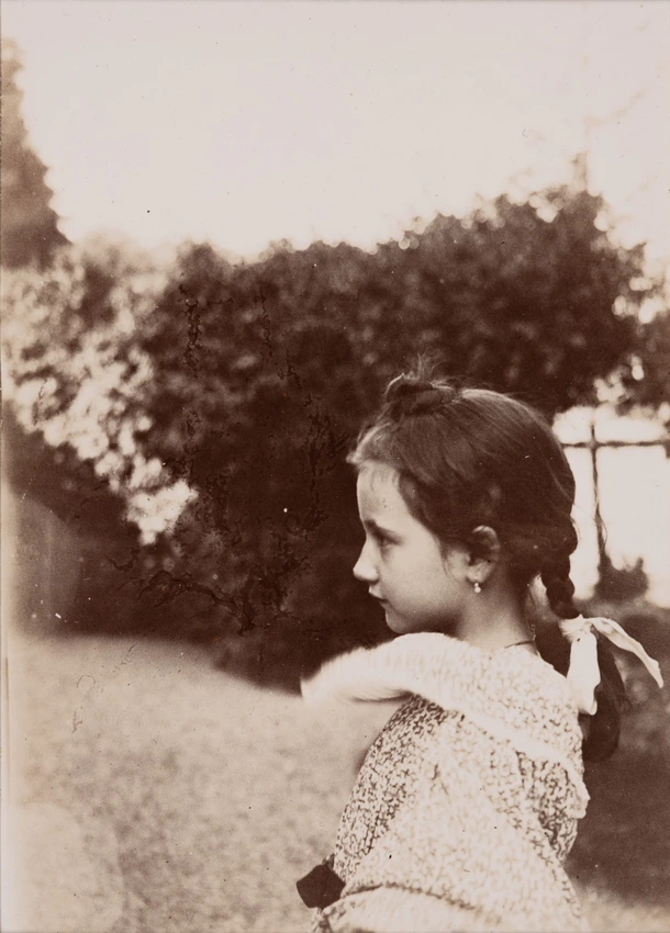 Denise de profil, coiffée avec une natte, se tenant à l'entrée d'une allée en graviers bordée d'arbres à droite - Emile Zola