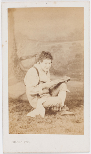 Franck - Paul Legrand en habit de scène (paysan), assis les jambes croisées, ten...