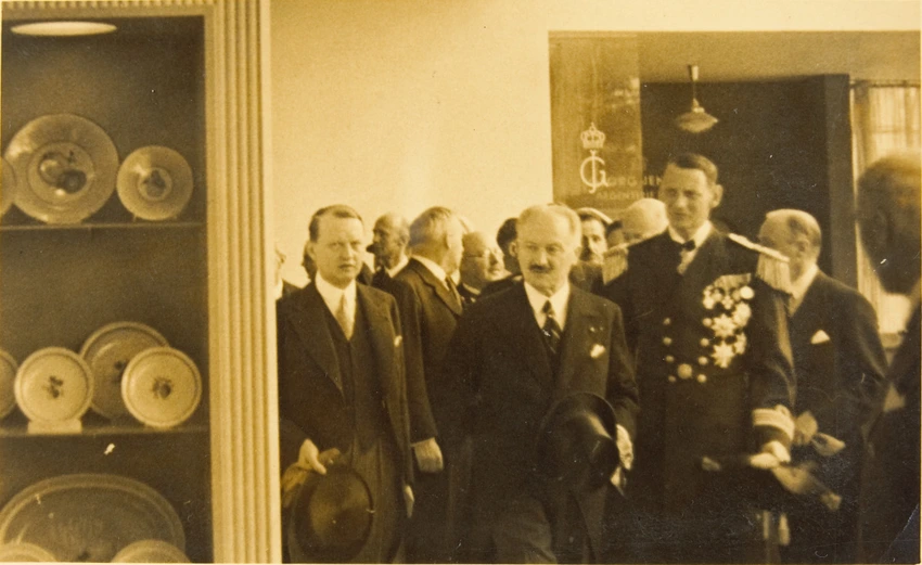 Groupe d'hommes debout dans une salle. Un homme porte des décorations sur sa veste. On voit à gauche de la photographie une étagère avec des céramiques - Anonyme