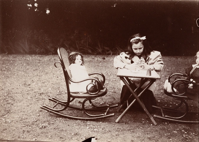 Au premier plan, décalé sur la droite, un ensemble de mobilier d’enfant est posé sur le sol du jardin : une petite table et deux fauteuils à bascule. Une poupée est assise dans chaque fauteuil. Denise est assise à la table, visage penché vers ses mains en train de manipuler des objets - Emile Zola